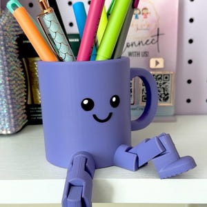 May include: A purple mug with legs and arms, shaped like a robot, holding a variety of colorful pens and markers. The mug has a happy face drawn on it.