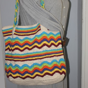 May include: A crocheted tote bag with a colorful striped and wavy pattern in shades of blue, green, yellow, orange, red, and purple. The bag has a long strap made of braided yarn in yellow and blue.