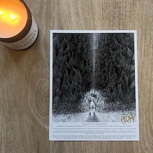 May include: A black and white print titled "Valley of Shadows" depicts a figure walking toward light in a dark cave. The print includes text at the bottom and is on a wooden surface. A lit candle sits to the left.
