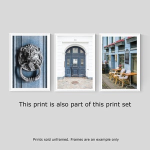 May include: A set of three framed prints featuring architectural details in a European city. The first print shows a close-up of a silver lion door knocker on a blue door. The second print shows a blue door with a white frame and the number 24 above it. The third print shows a street scene with blue buildings and tables and chairs set up outside a cafe.