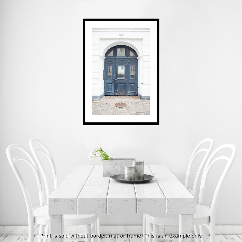 May include: A photograph of a blue door with a white frame and a white building behind it. The door has a rounded archway and a metal door handle. The number 24 is visible above the door.