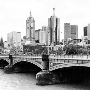 May include: A black and white photograph of a bridge over a river with a city skyline in the background. The bridge is made of stone and has a series of arches. The city skyline is made up of tall buildings, including a church steeple.