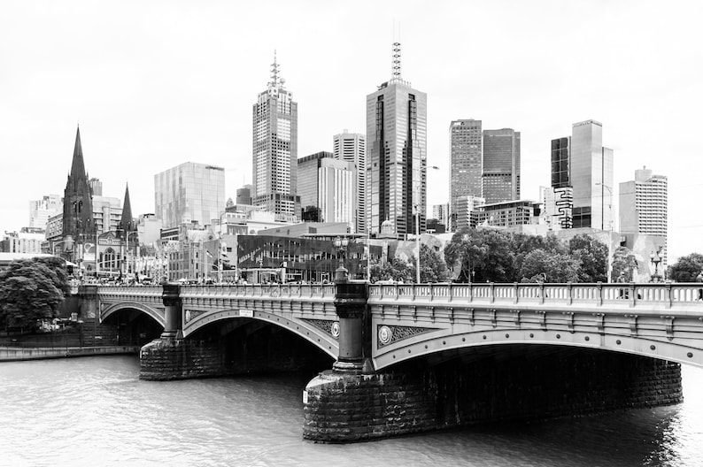 May include: A black and white photograph of a bridge over a river with a city skyline in the background. The bridge is made of stone and has a series of arches. The city skyline is made up of tall buildings, including a church steeple.