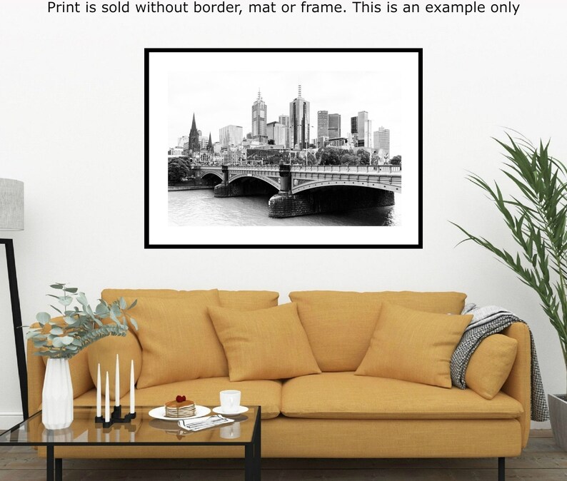 May include: Black and white photograph of a city skyline with a bridge in the foreground. The bridge has multiple arches and is made of stone. The city skyline is in the background and includes tall buildings. The photo is framed in a black frame.