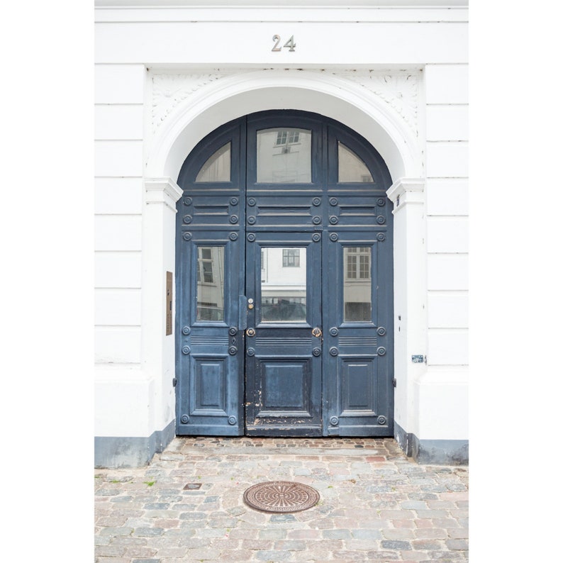 May include: A blue wooden door with a decorative archway and the number 24 above it. The door has multiple panels and is set in a white wall.