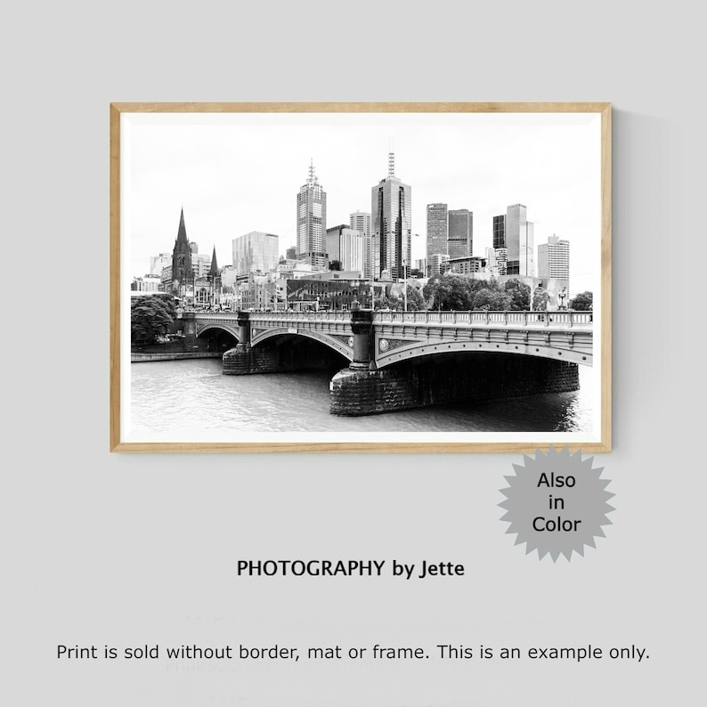 May include: Black and white photograph of a city skyline with a bridge in the foreground. The city skyline features tall buildings and a river. The bridge is a stone arch bridge with a road running over it. The photograph is framed in a wooden frame.