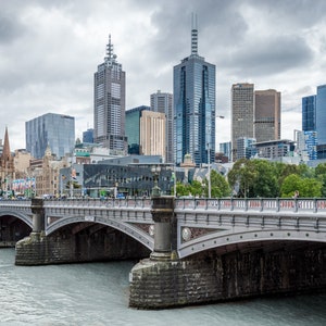 May include: A gray stone bridge with arches over a river in front of a city skyline. The city skyline includes several tall buildings, including a church with a spire.