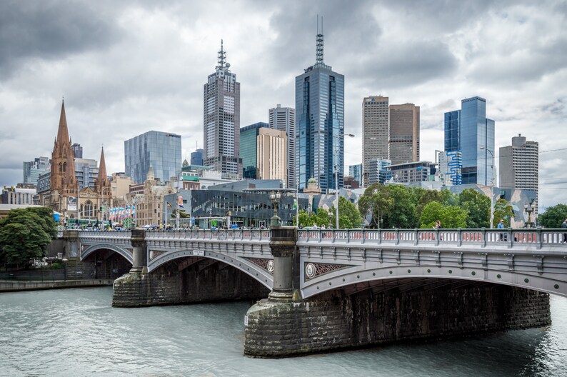 May include: A gray stone bridge with arches over a river in front of a city skyline. The city skyline includes several tall buildings, including a church with a spire.