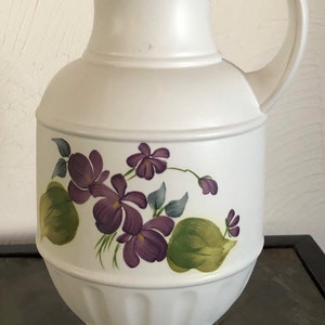 May include: White ceramic pitcher with a floral design featuring purple violets and green leaves. The pitcher has a wide base and a handle.