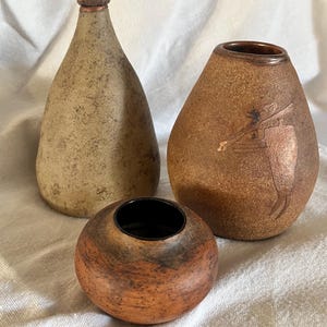 Peut inclure: Trois vases en céramique faits à la main dans des tons terreux. Le vase le plus haut est beige clair avec une surface texturée. Le vase moyen est marron avec un motif de silhouette. Le plus petit vase est rond et marron moucheté.