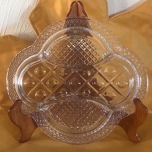 May include: A clear, divided glass serving dish with a diamond and dot pattern. The dish is supported by a wooden stand, and has a decorative, scalloped edge. The dish is designed to hold multiple items.