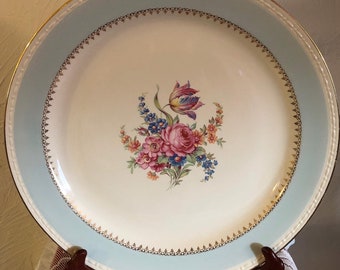 May include: A vintage porcelain dinner plate with a light blue rim and a gold trim. The center of the plate features a floral design with pink, yellow, and blue flowers.