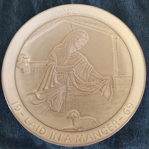 May include: A beige ceramic plate with a raised design of a woman holding a baby in a manger. The plate has the text "19-LAID IN A MANGER-69" around the edge.