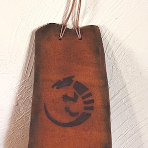 May include: A rectangular, brown, leather-like wall hanging with a dark lizard design. The hanging is suspended by a brown cord attached to a decorative, dark brown button. The background is a white wall.