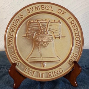 May include: A tan ceramic plate with a brown rim and a raised image of the Liberty Bell. The plate has the text "America's Symbol of Freedom 1776 Let It Ring 1976" around the rim.