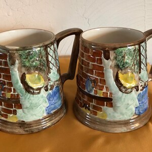 May include: Two ceramic mugs with brown, red, green, blue, and yellow painted designs. The mugs feature a brick wall and a person in a green and blue outfit.