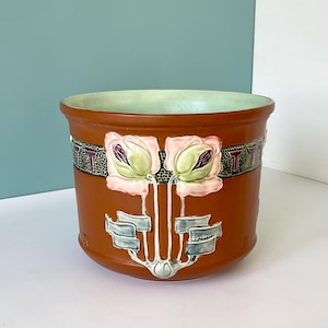 Könnte beinhalten: Ein brauner Keramik-Blumentopf mit hellgrünem Innenraum. Der Topf hat ein dekoratives Band mit dem Buchstaben "T" und Blumendesigns mit rosa und grünen Blumen. Der Sockel hat ein stilisiertes Design.