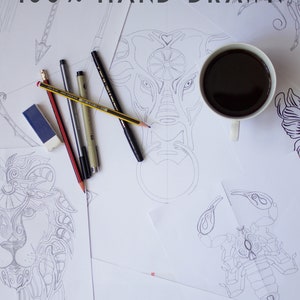 May include: A flat lay image of a white coffee mug filled with black coffee, a pencil, and several black pens on a white surface with several hand-drawn designs. The designs include a lion, a scorpion, and a bull's head. The text "100% HAND DRAWN" is written at the top of the image.