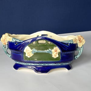 Könnte beinhalten: Ein Keramik-Pflanzgefäß in tiefblau, mit einem rechteckigen grünen Feld und floralen Akzenten. Das Pflanzgefäß hat einen cremefarbenen Innenraum und ist mit stilisierten floralen Griffen verziert. Der Sockel hat eine dekorative Zierleiste.