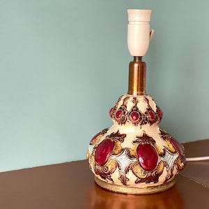 Puede incluir: Una lámpara de mesa vintage con base crema, adornada con detalles ovalados burdeos y patrones decorativos dorados y marrones. La lámpara tiene un cuello de latón y un portalámparas blanco. La lámpara está enchufada a una toma de corriente.