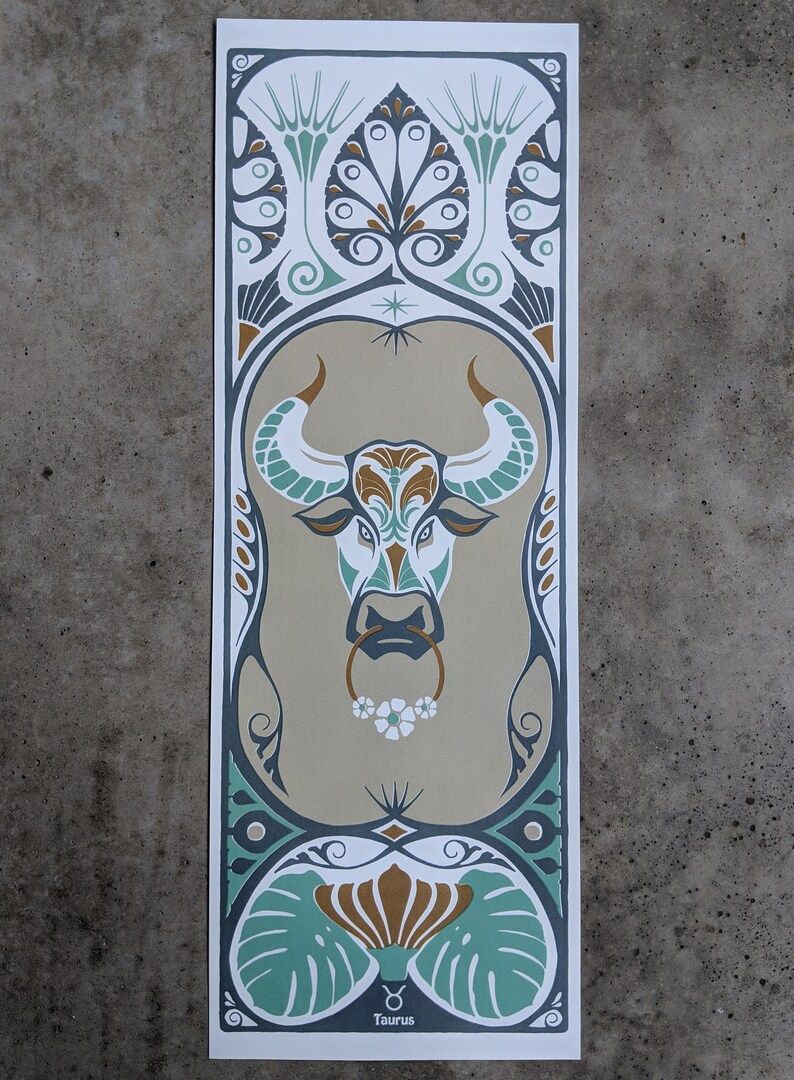 May include: A stylized illustration of the Taurus zodiac sign, featuring a bull's head with a floral nose ring. The bull is surrounded by intricate floral patterns in shades of green, gold, and brown. The text "Taurus" is printed at the bottom of the image.