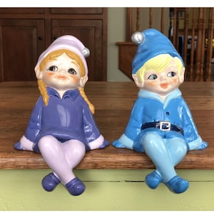May include: Two ceramic elf figurines, one in blue and one in purple, sitting on a wooden surface. Both elves have blonde hair, blue eyes, and are wearing hats.