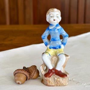 May include: A small porcelain figurine of a boy in a blue shirt with yellow shorts and red shoes, seated on a tree stump. Two acorns are next to the figurine. The figurine is painted with fine details.