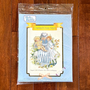May include: A counted cross stitch pattern featuring a mother and two children sitting on a bench. The pattern is titled "Hopes and dreams are life's surbeams" and is from Holly Hobbie. The kit includes 14 count Aida cloth, ribbon, needle, chart, and easy instructions.