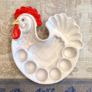 May include: A white ceramic serving tray shaped like a rooster with a bright red comb and wattle, a yellow beak, and six circular indentations along the bottom edge for holding items, featuring a glossy finish and molded feather details.