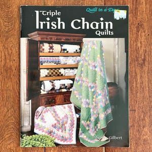 May include: A book titled "Triple Irish Chain Quilts" with a photo of quilts and a wooden cabinet. The cover features the title in large white letters. A green quilt is draped over a wooden chest. Several quilts are visible inside the cabinet.