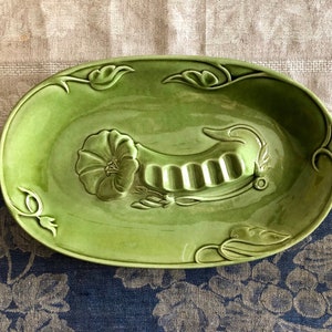 May include: A green ceramic ashtray with a floral design and a built-in cigarette rest. The ashtray is oval shaped and has a raised rim.