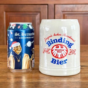 May include: A blue St. Bernard Christmas Ale can next to a Binding Bier stein. The can features an illustration of a saint wearing a Santa hat. The stein is cream-colored with the Binding Bier logo and text.