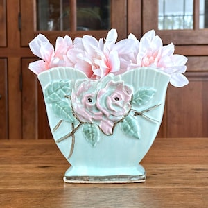 May include: A light blue ceramic vase with a rose design and gold trim, filled with pink and white flowers. The vase features embossed roses and leaves. The vase is sitting on a wooden surface.