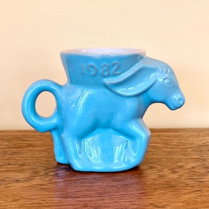 May include: A turquoise ceramic mug shaped like a donkey, with the year "1982" embossed on the rim. The mug has a rounded handle and a white interior, set against a wooden surface.