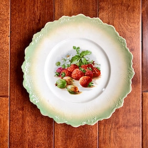 May include: A white ceramic plate with a green rim features a painted design of red strawberries and green gooseberries on a vine. The plate has a scalloped edge.