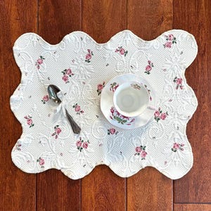 May include: A white, scalloped placemat with a quilted floral pattern featuring pink roses. A teacup and saucer with matching rose details sit on the mat, along with a silver spoon. The scene evokes a vintage tea party aesthetic.