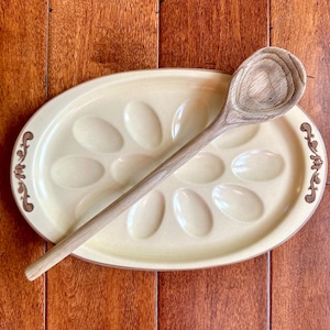 May include: A light yellow ceramic egg plate with brown floral accents and a wooden spoon. The plate has six indentations for eggs.