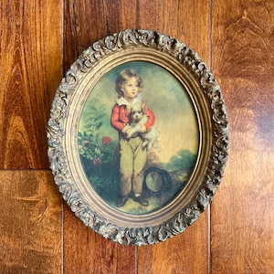 May include: Oval framed print featuring a young child in a red jacket and tan pants holding a small white dog. The print has a gold ornate frame and is set against a landscape background with flowers and a hat on the ground.