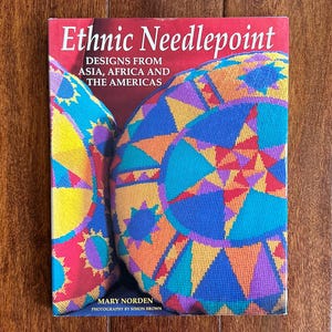 May include: A book titled "Ethnic Needlepoint" with a red cover. The cover features colorful needlepoint designs in blue, orange, yellow, and purple. The text reads "DESIGNS FROM ASIA, AFRICA AND THE AMERICAS" and the author is Mary Norden.
