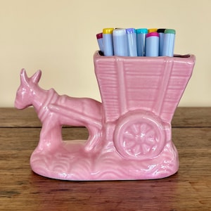 May include: A pink ceramic donkey pulling a pink wagon with a wooden base. The wagon is filled with colorful markers.