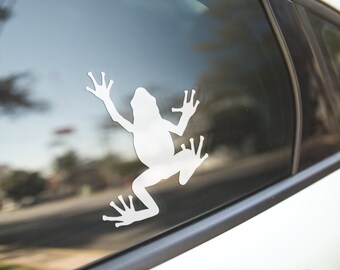 Puede incluir: Una calcomanía de silueta de rana blanca en la ventana de un coche. La calcomanía es un simple contorno de una rana con las patas extendidas.