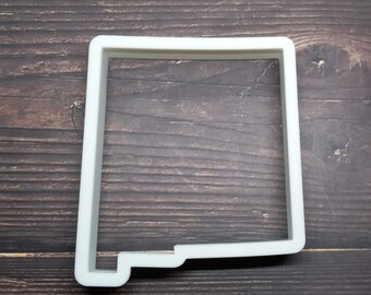 May include: A white plastic cookie cutter in the shape of the state of New Mexico. The cutter has a rectangular shape with a small notch on the bottom left side. The cutter is made of a thick, durable plastic.
