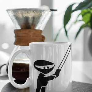 Puede incluir: Taza de cer&aacute;mica blanca con un ninja negro sosteniendo una espada. La taza est&aacute; sobre un posavasos de pizarra negra.
