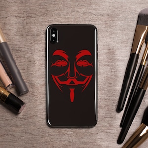 May include: A black phone case with a red Anonymous mask graphic. The phone case is on a wooden surface with makeup brushes.