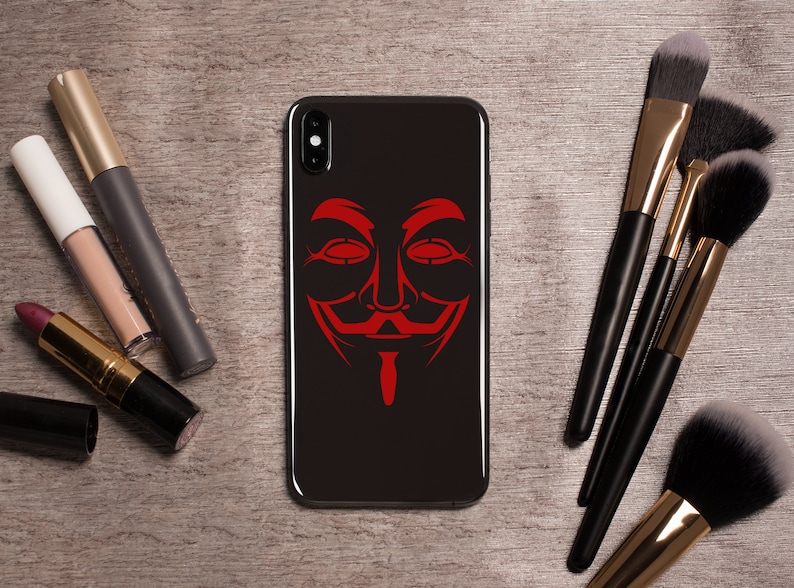 May include: A black phone case with a red Anonymous mask graphic. The phone case is on a wooden surface with makeup brushes.
