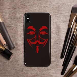Puede incluir: Una funda de tel&eacute;fono negra con un gr&aacute;fico de m&aacute;scara roja de Anonymous. La funda del tel&eacute;fono est&aacute; sobre una superficie de madera con pinceles de maquillaje.