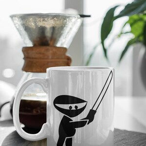 May include: White ceramic mug with a black ninja holding a sword graphic. The mug is sitting on a black slate coaster.