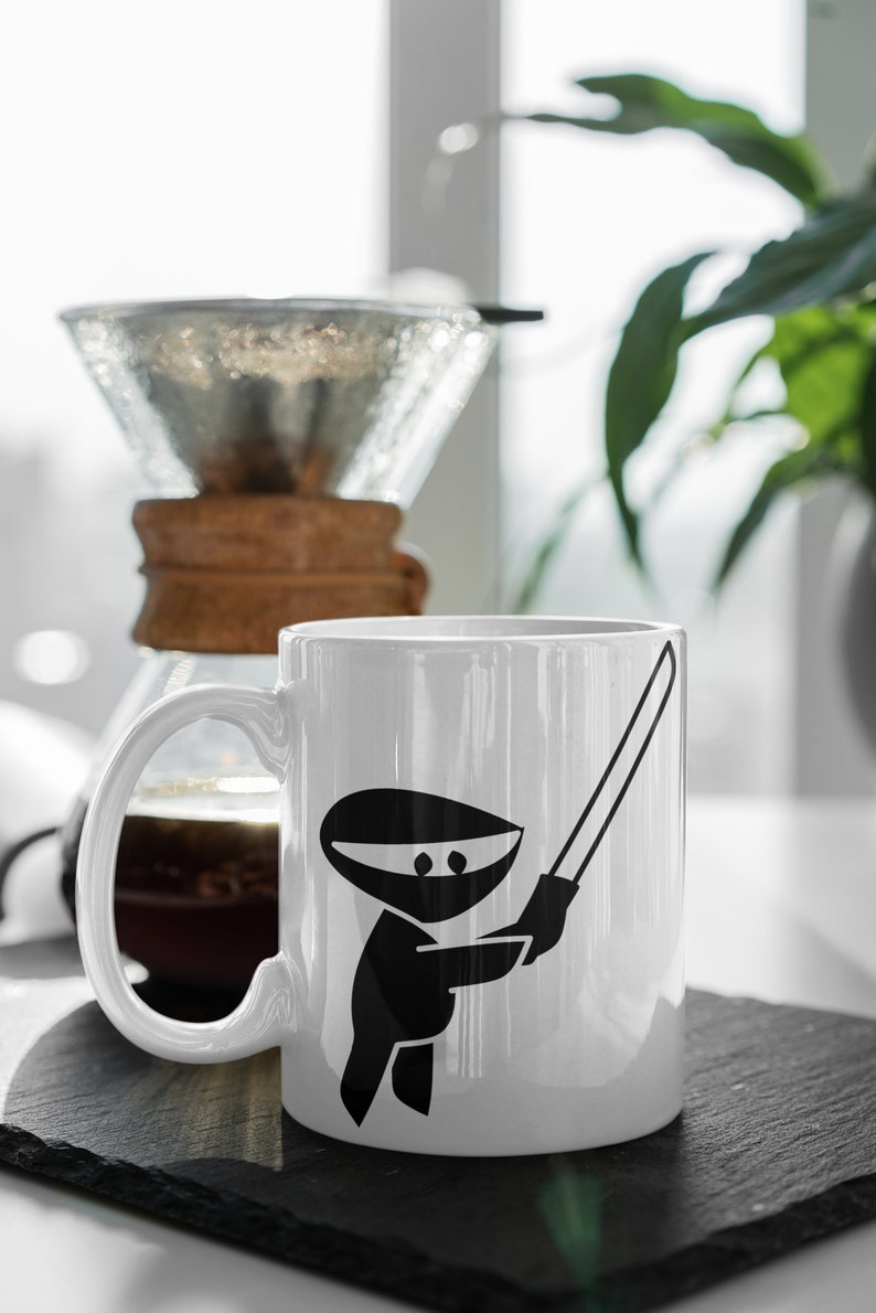 May include: White ceramic mug with a black ninja holding a sword graphic. The mug is sitting on a black slate coaster.