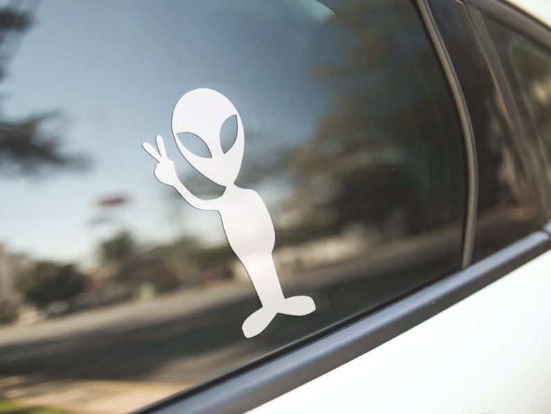 Puede incluir: Una pegatina de extraterrestre blanca con un signo de paz en la ventana de un coche.