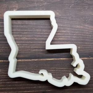 May include: A white plastic cookie cutter in the shape of the state of Louisiana. The cutter has a simple, clean design, with a uniform thickness. The state's outline is clearly defined, with its distinctive shape.